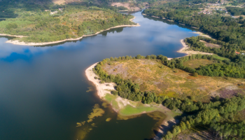 A wide aerial view of a large lake surrounded by green woodland and rolling hills. The shoreline curves around several inlets, with patches of sandy or rocky banks visible along the water’s edge. The foreground shows a small peninsula covered in low vegetation, leading into calm, reflective blue water. In the distance, more forested hills and a small cluster of buildings are visible.