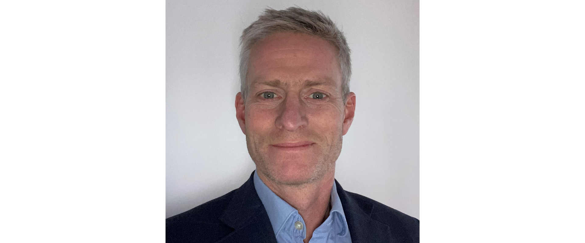 Head-and-shoulders professional portrait of a person with short light-colored hair, wearing a light blue button-down shirt and a dark navy blazer. The person is facing the camera against a plain light gray wall, with even lighting and a neutral background. The image is vertically framed and resembles a corporate or business profile photograph.