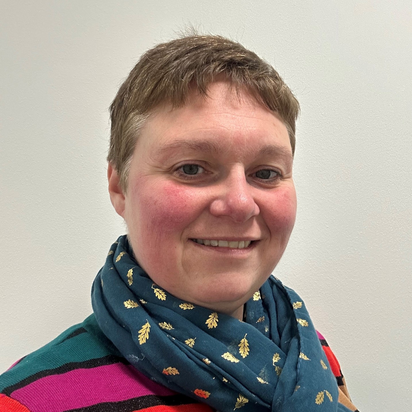 white woman with short grey hair, rosey cheeks and wearing a rainbow striped jumper and blue neck scarf