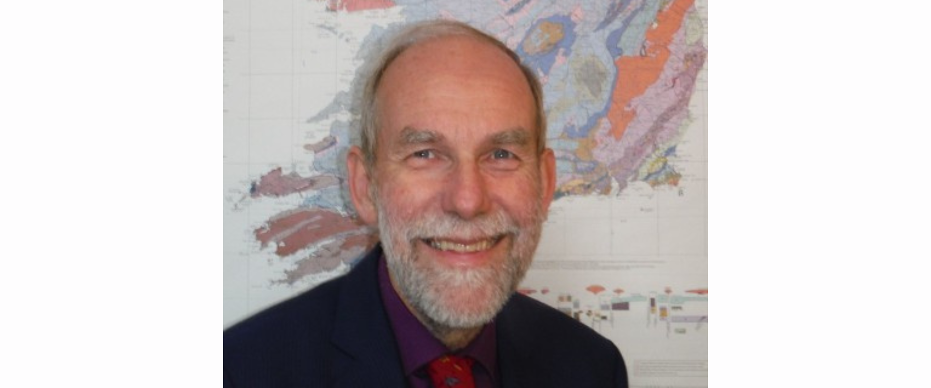  Person wearing a dark jacket, shirt, and patterned tie, standing in front of a large, colorful geological map mounted on a wall.