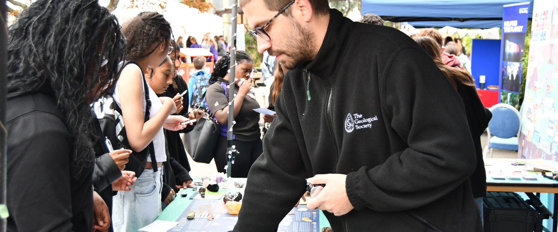 a man with glasses and a black jacket talking to a black woman and pointing to a display of rocks on a table