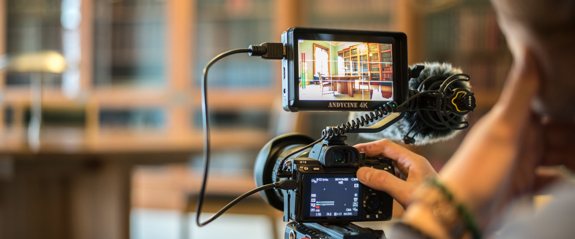 A person recording a video in a library, surrounded by books and shelves, focused on their camera.