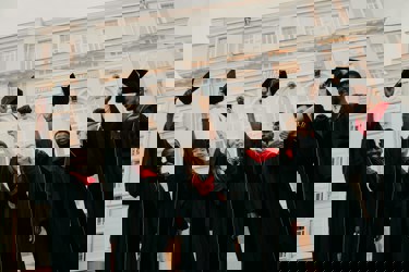 A group of graduates in black gowns and red hoods joyfully raises their caps in front of a university building.