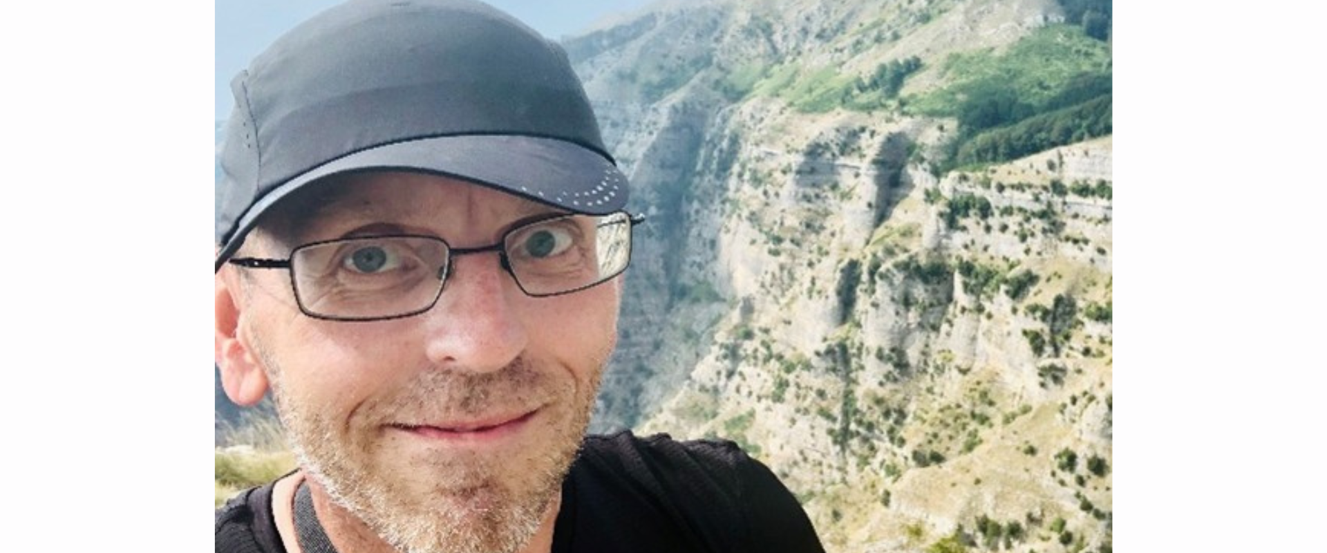 A person wearing a dark cap and backpack standing outdoors with a rugged, mountainous landscape in the background. The scene shows steep rocky cliffs, patches of vegetation, and distant tree‑covered slopes under a bright sky.
