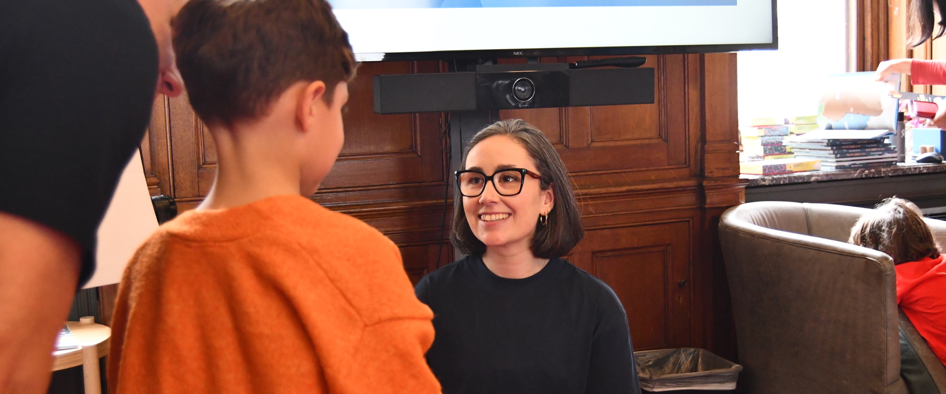 woman with glasses and short bobbed hair smiling at a small child with an adult next to them