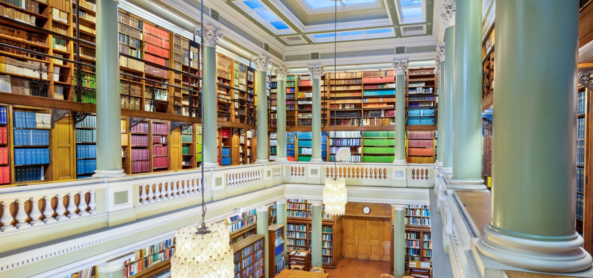 The Upper Library of the Geological Society of London, taken from an upstairs gallery looking down at table and chairs on a maroon carpet surrounded by book-lined shelves. Two bright glass chandeliers hang above the library.