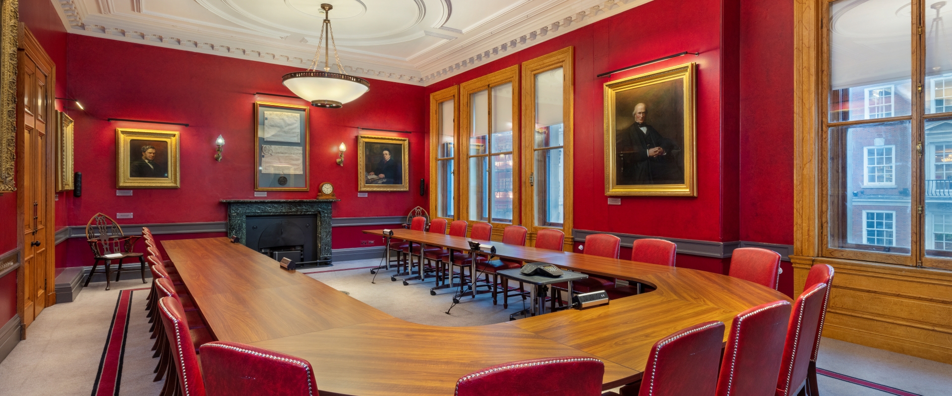 Boardroom with a long wooden table, red walls adorned with portraits, and large windows, creating a formal and historic atmosphere.