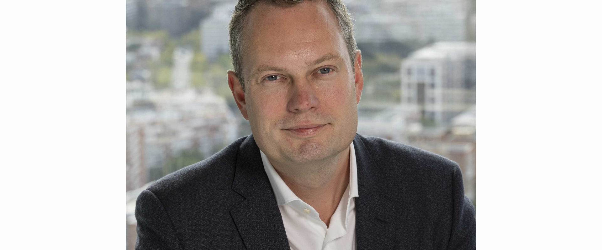 A person wearing a dark textured blazer over a white collared shirt is seated indoors with a cityscape visible through large windows in the background. The skyline includes tall buildings, residential blocks, and soft, diffused daylight. The individual is positioned centrally, shown from the chest up.