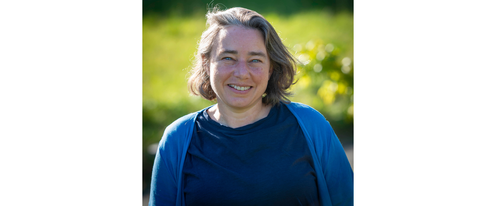 Outdoor portrait of a person standing in front of a softly blurred green landscape. The person has shoulder-length hair and is wearing a dark blue top with a lighter blue cardigan. The image is lit by bright natural sunlight, with the subject centered in the frame and the background consisting of green vegetation and foliage rendered out of focus. No visible text appears in the image.