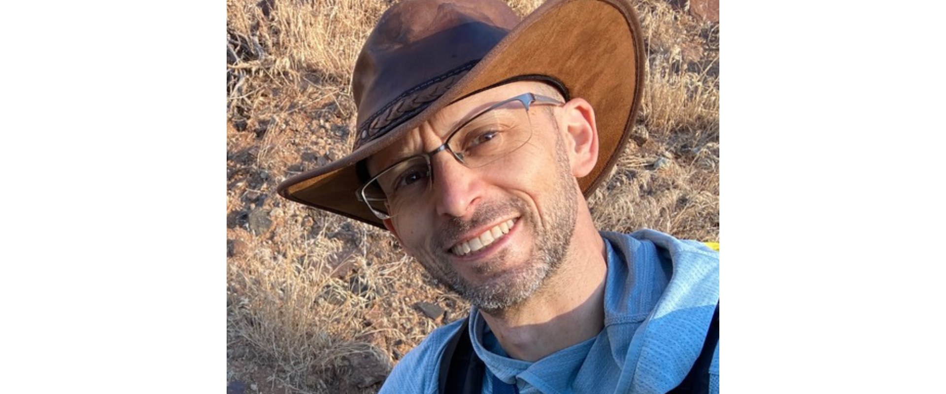 Close-up outdoor portrait of a person wearing a wide-brimmed brown hat, glasses, and a light blue hooded top. The person is positioned in the foreground, angled slightly toward the camera. The background shows a dry, rocky landscape with tan grasses and scattered stones, illuminated by warm natural sunlight. No visible text appears in the image.