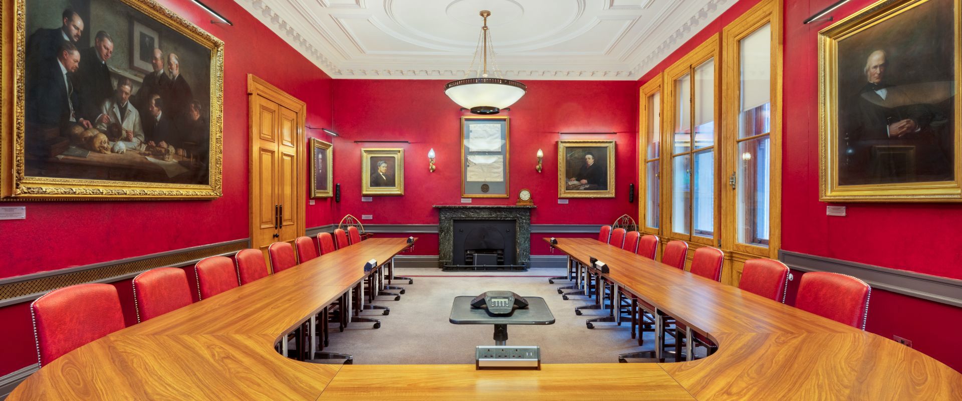 Boardroom with a long wooden table, red walls adorned with portraits, and large windows, creating a formal and historic atmosphere.