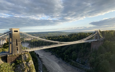 A panoramic view of the Clifton Suspension Bridge in Bristol, spanning a gorge with lush greenery and dramatic clouds overhead.
