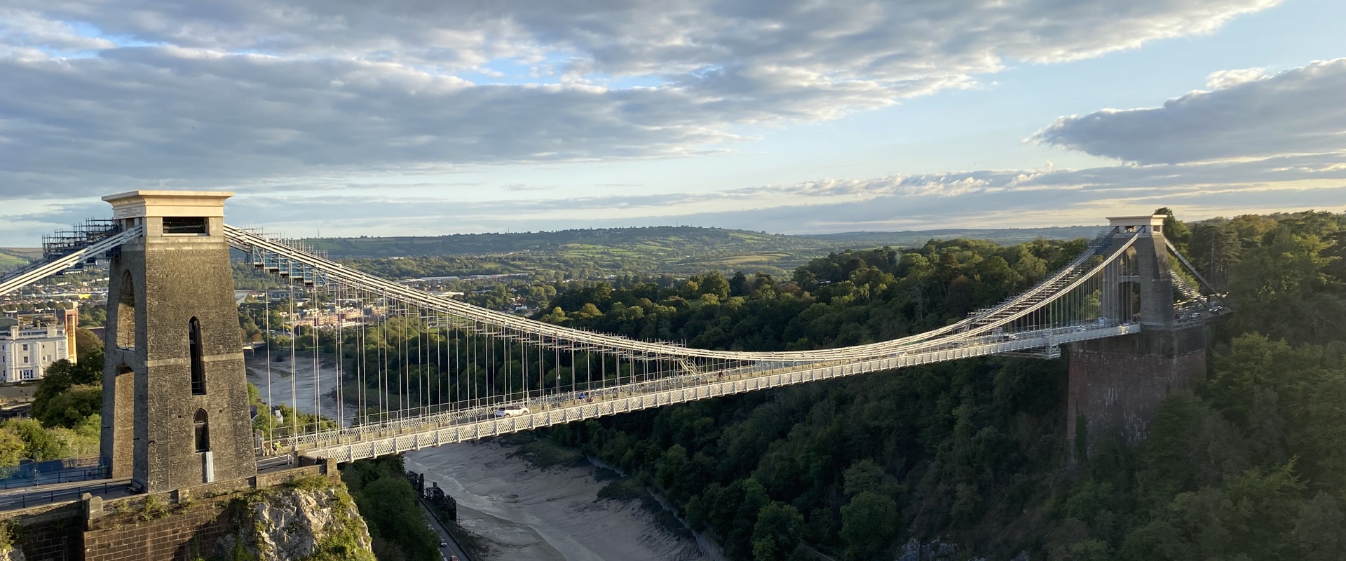 A panoramic view of the Clifton Suspension Bridge in Bristol, spanning a gorge with lush greenery and dramatic clouds overhead.