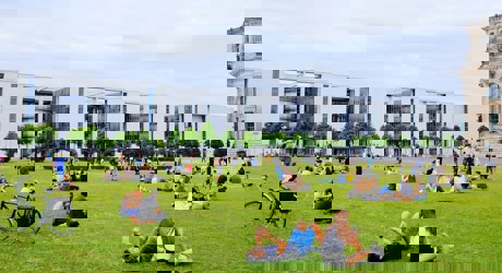 People relax on the lawn in front of modern buildings, enjoying a lively day in a public space filled with greenery.