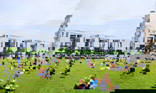 People relax on the lawn in front of modern buildings, enjoying a lively day in a public space filled with greenery.