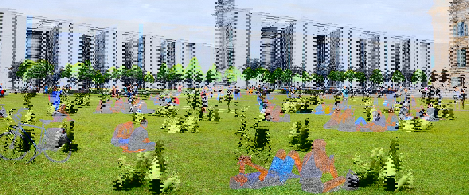 People relax on the lawn in front of modern buildings, enjoying a lively day in a public space filled with greenery.