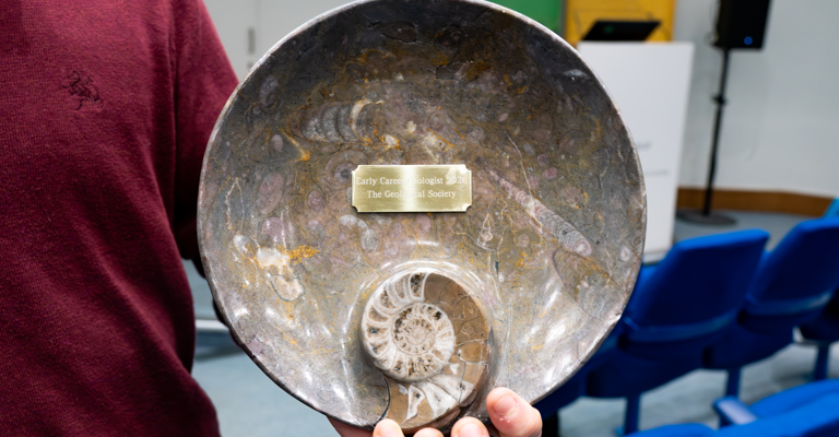 A hand holding a concentric trophy with an ammonite and a plaque