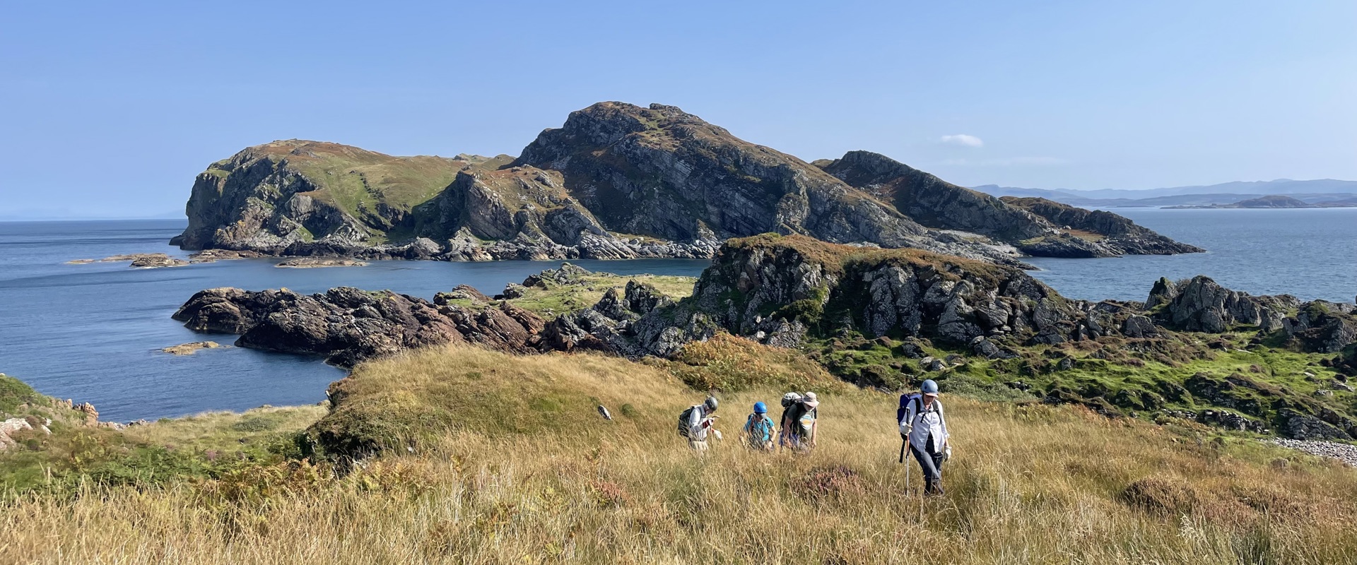 Five hikers traverse a grassy coastal landscape with rocky hills and a calm sea under a clear blue sky, conveying a sense of adventure and tranquility.