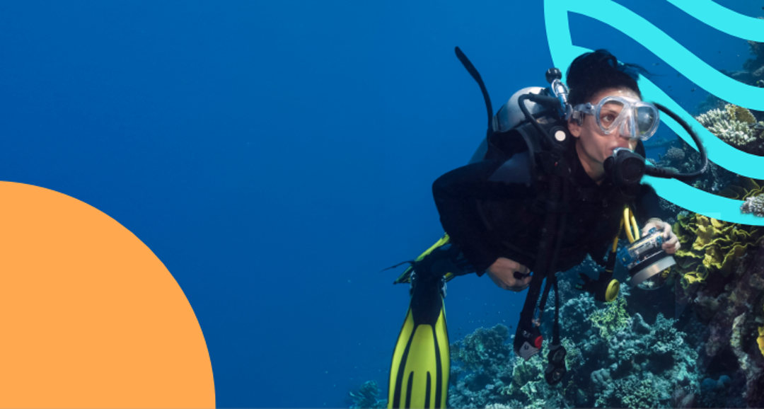 A diver is underwater near vibrant corals, equipped with scuba gear and a camera, surrounded by a deep blue ocean