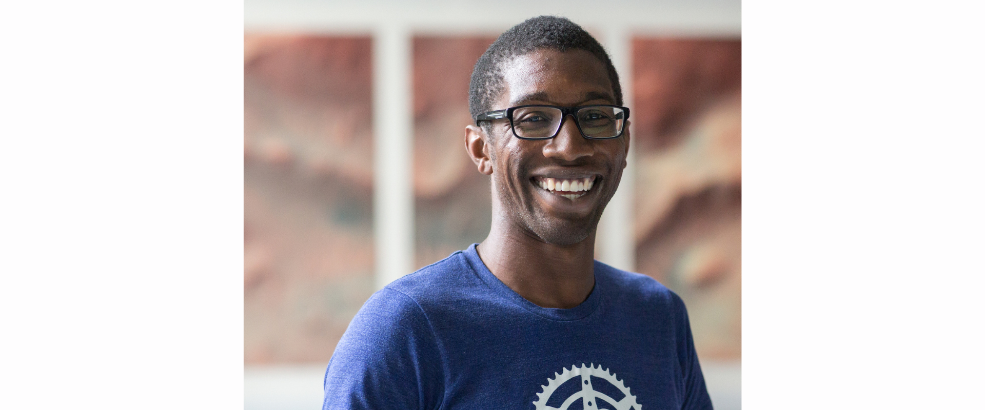 A person wearing a blue T‑shirt with a circular gear‑shaped graphic stands indoors in front of a softly focused background featuring large abstract artworks in warm earth tones.