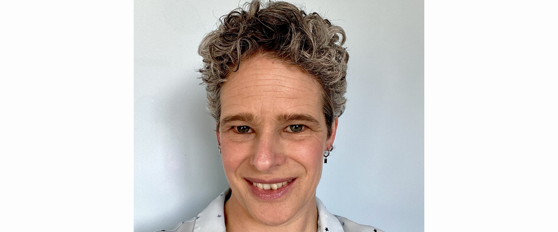 A person with short, curly hair is photographed from the shoulders up against a plain light‑coloured wall. The person is wearing a light shirt with a subtle star pattern.