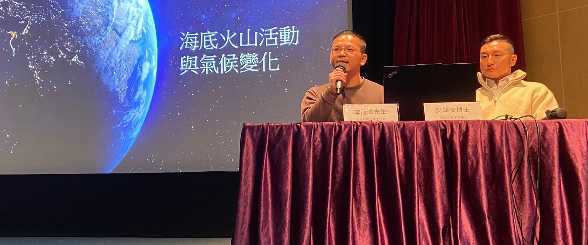 two men one holding a mic sat at a table with a projector screen behind them
