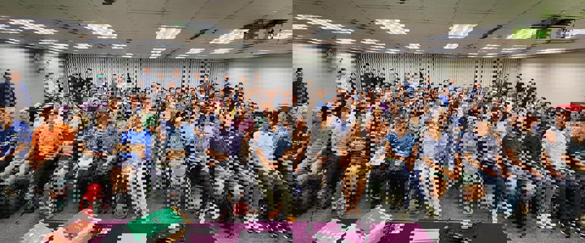 A large group of attendees gathers in an indoor seminar space, with tables displaying papers and electronic devices in the foreground.