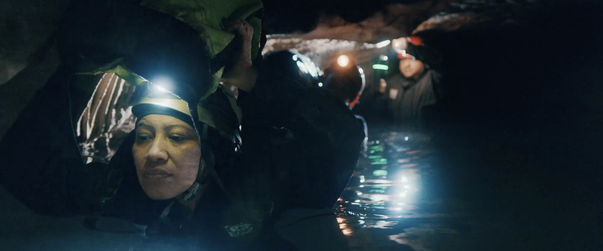 Woman and man passing through a narrow, flooded cave passage with bags over their heads and water up until their necks