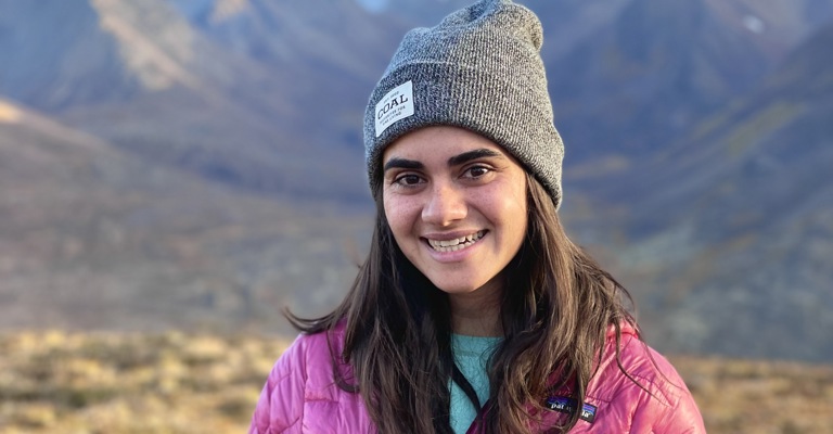 young woman with long brown hair with a beanie and pink jacket stood in front of a mountain landscape