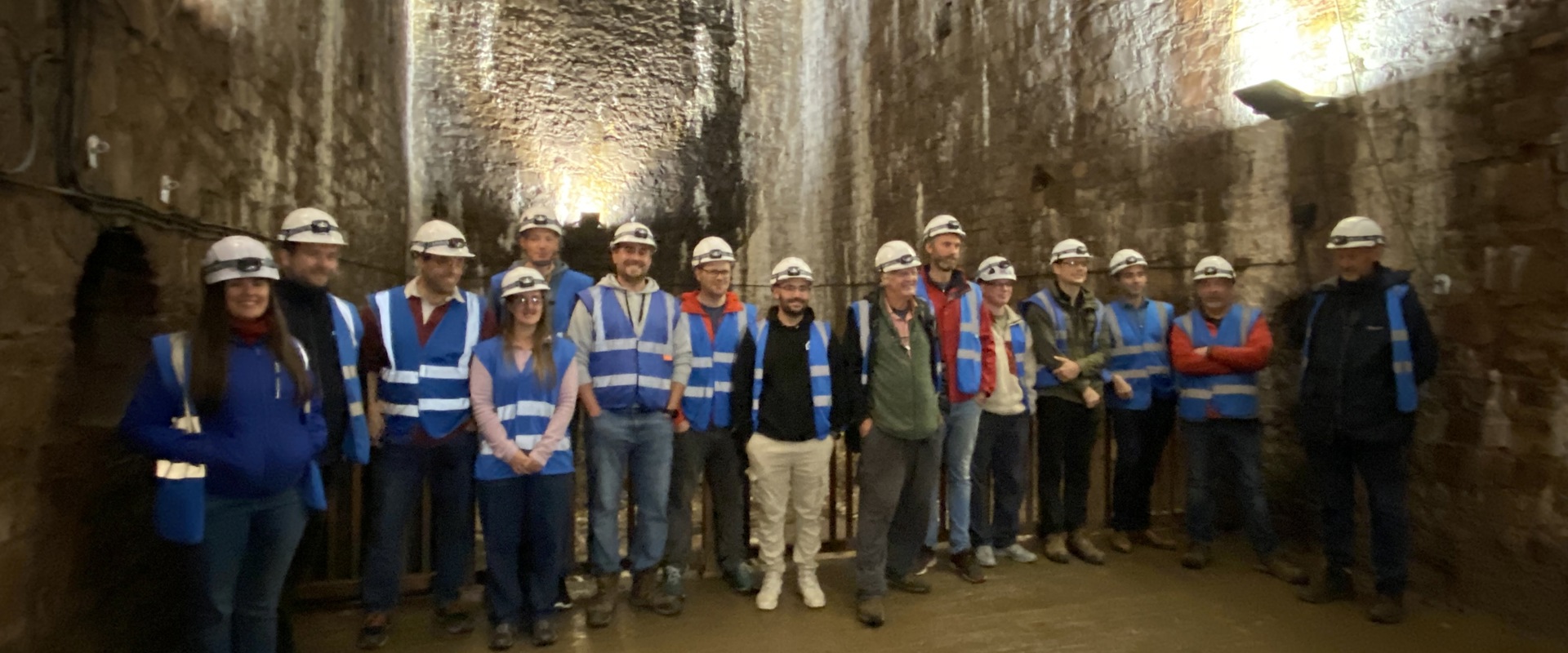 A large group of people wearing safety helmets and blue vests stands indoors, surrounded by tall, illuminated stone walls.