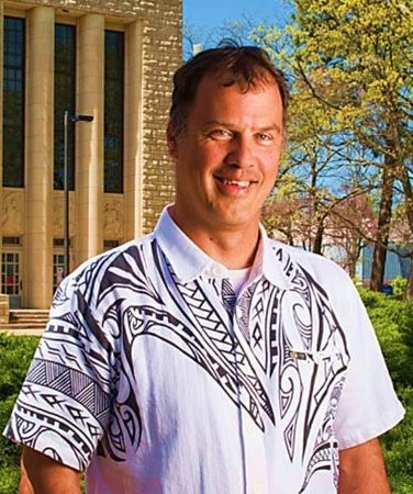 white man with brown hair and a white short sleeve shirt with a black pattern on it stood in front of a building with trees to one side
