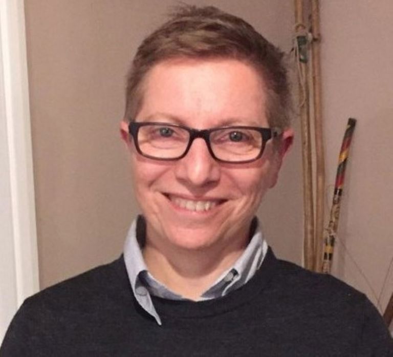 white woman with short hair and glasses wearing a grey jumper with a shirt underneath