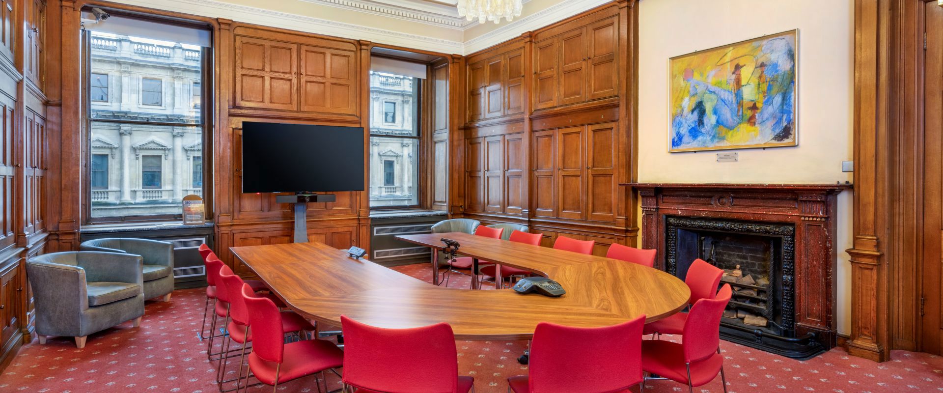 Modern conference room featuring a large oval table, red chairs, a TV, and wooden paneling, with large windows and artwork.