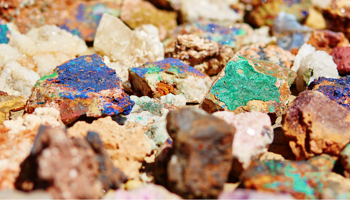 Assorted colorful mineral and rock specimens, featuring vivid blue and green mineral coatings and white crystals, photographed in bright light with a blurred background.