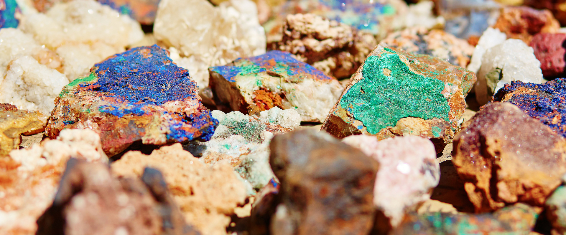 Assorted colorful mineral and rock specimens, featuring vivid blue and green mineral coatings and white crystals, photographed in bright light with a blurred background.