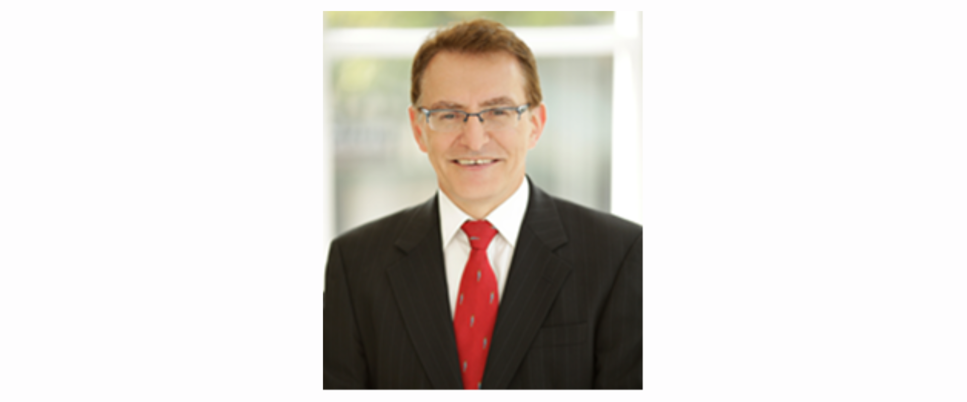 A person wearing a dark business suit with a white shirt and a red patterned tie, standing indoors in front of a softly lit background with light-coloured panels.