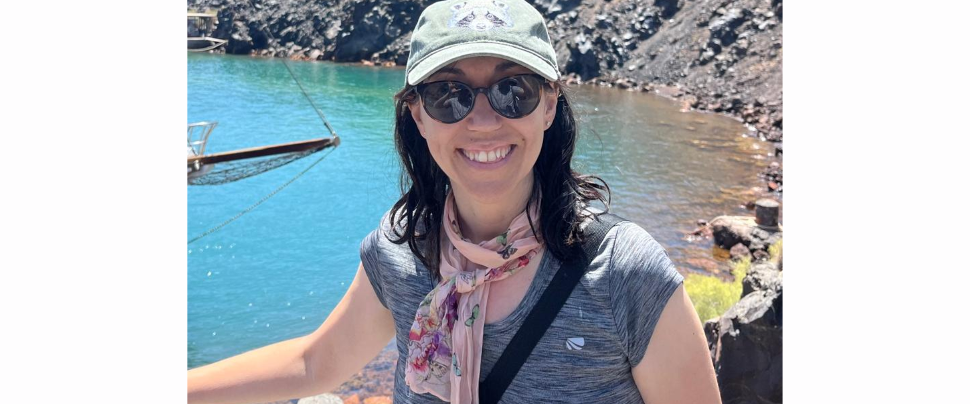 A person wearing a light green cap, dark round sunglasses, and a grey short-sleeve top stands in front of a bright blue body of water surrounded by rocky terrain. A light scarf is tied around the neck, and a black strap crosses the chest. A rope railing runs across the foreground. In the background, there are dark rocks and a small structure or platform extending toward the water. The scene is brightly lit, suggesting a sunny outdoor location.