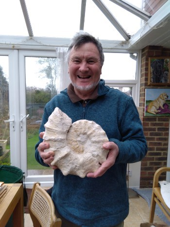 White man with grey hair and grey beard smiling holding a large ammonite fossil, stood inside a house with doors and a garden behind him