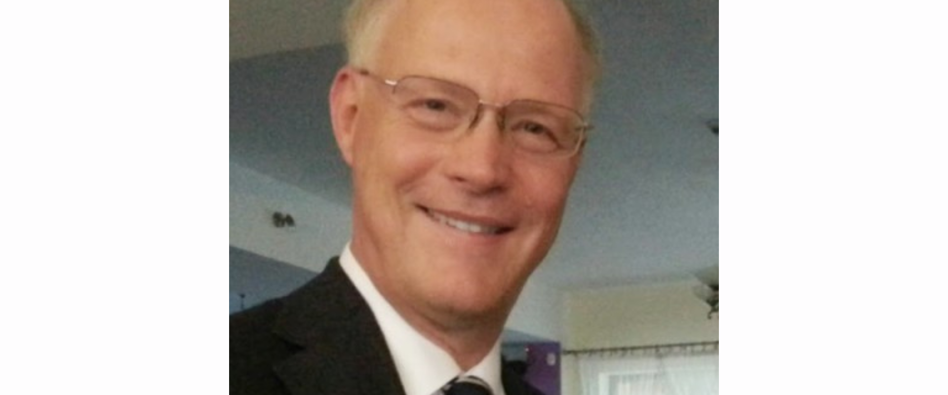Portrait of a person wearing glasses, a dark suit jacket, a white shirt, and a striped tie, photographed indoors against a softly lit background with a window and light‑coloured walls.