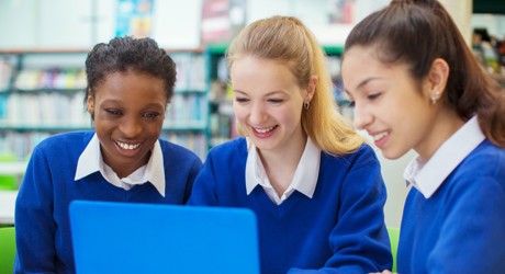 Two white girls and one black girl all in school uniform of blue jumpers and white blouses, all smiling looking at a laptop