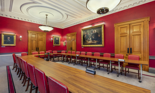 Boardroom with a long wooden table, red walls adorned with portraits, and large windows, creating a formal and historic atmosphere.