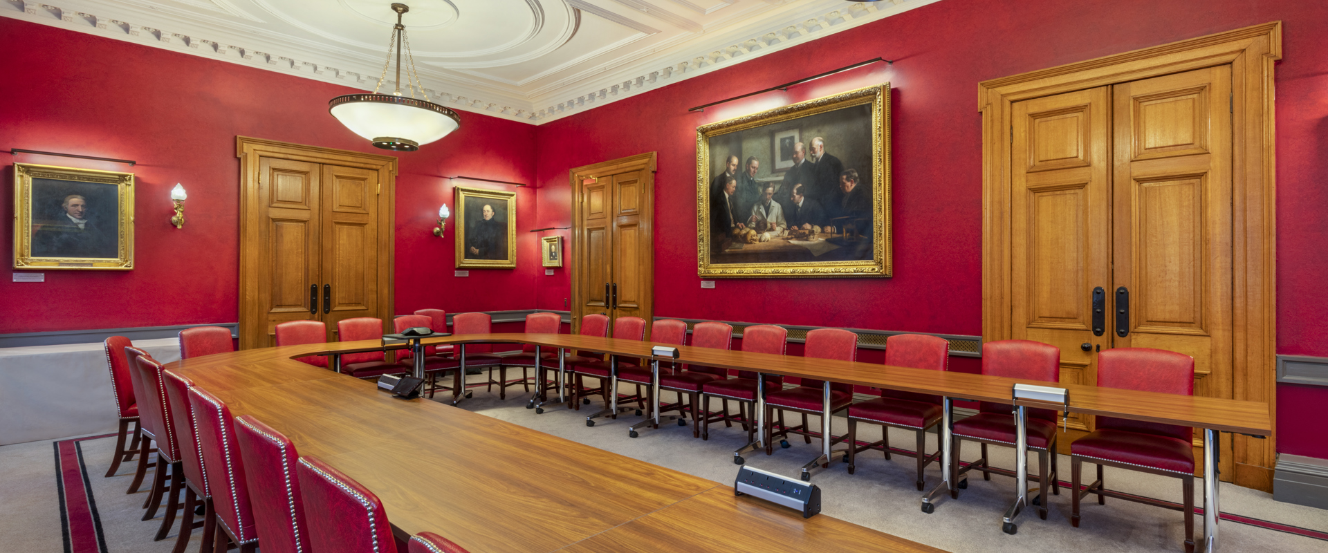 Boardroom with a long wooden table, red walls adorned with portraits, and large windows, creating a formal and historic atmosphere.