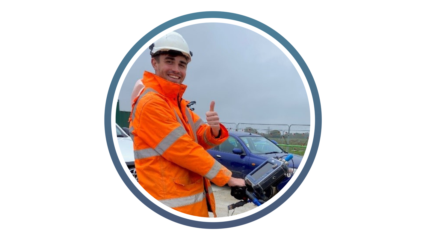 young man in high vis jacket and white hard hat stood in front of a blue car and holding electronic device