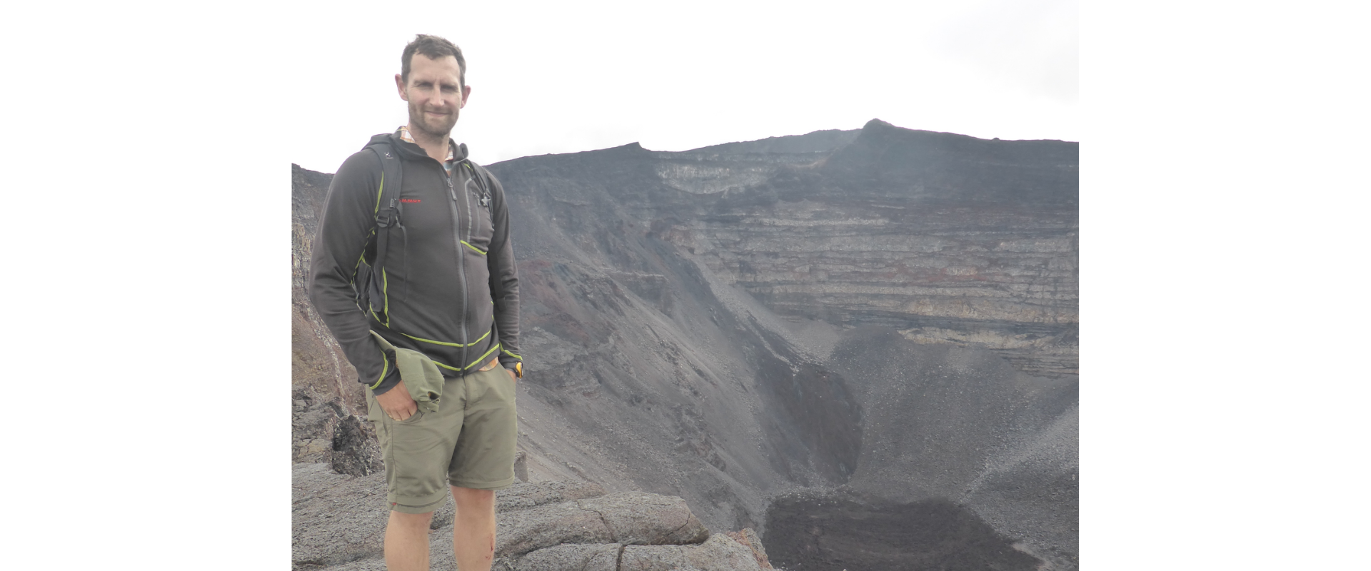 Full-body outdoor photograph of a person standing on a rocky ledge at the edge of a large volcanic crater. The person is wearing hiking boots, shorts, a dark zip-up jacket, and a backpack. Behind them, steep crater walls display layered rock formations descending into a dark, rugged basin. The landscape is predominantly grey and rocky, with an overcast sky creating soft, diffused lighting. No visible text appears in the image.