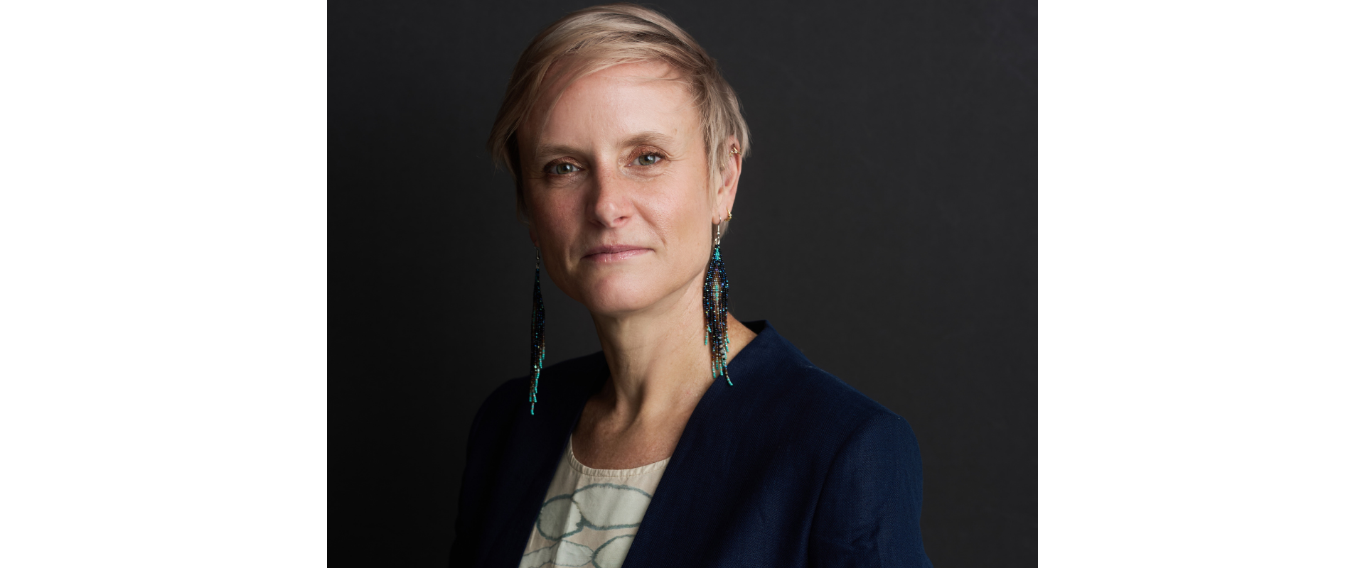 Professional studio portrait of a person with short light-colored hair, wearing a dark blue blazer over a light patterned top. The person faces the camera against a dark, softly textured background and wears long beaded earrings. Soft side lighting highlights the face and upper body, creating a formal and polished headshot.