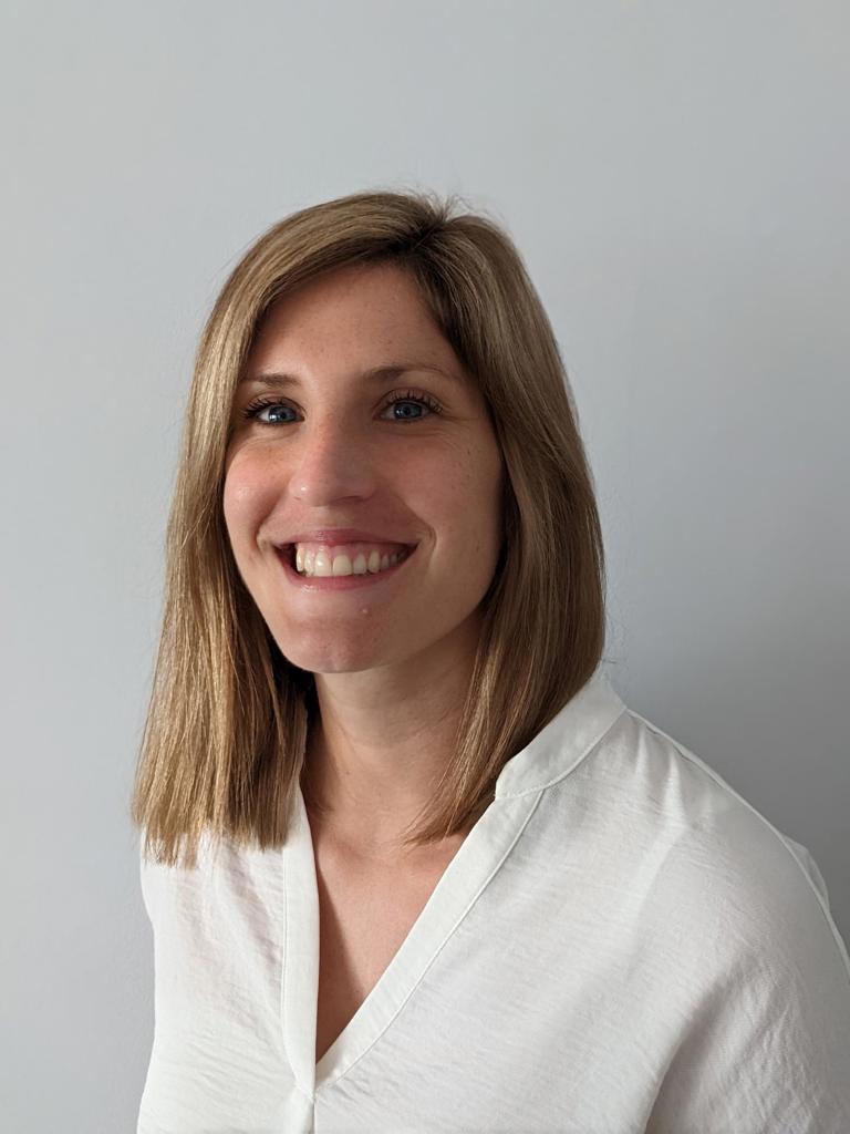 A woman with shoulder-length blonde hair smiling in a white collared shirt against a grey background.