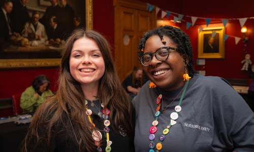 Two women stand together, showcasing colorful badge necklaces, with a historic painting in the background and festive decorations.