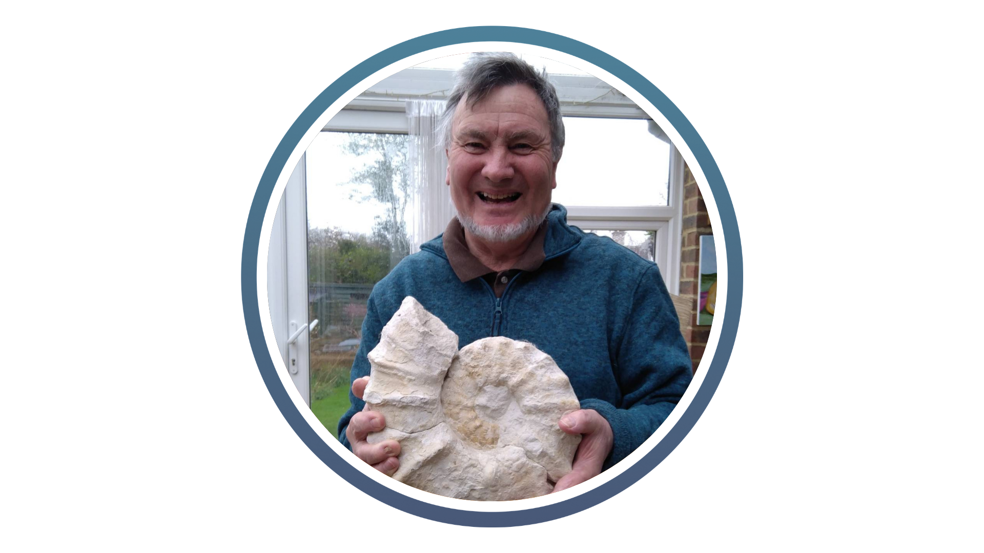 White man with grey hair and grey beard smiling holding a large ammonite fossil, stood inside a house with doors and a garden behind him