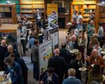 A lively networking event in a library, with attendees enjoying food and discussing onlookers among colorful posters and bookshelves.