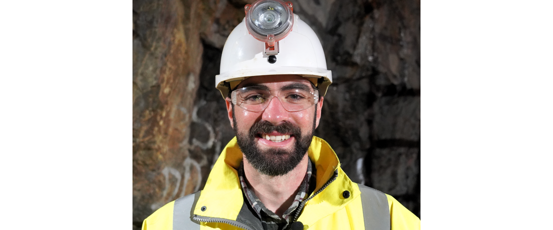 A person stands in a rocky underground space, wearing a white hard hat with a headlamp, clear safety goggles, and a bright yellow high‑visibility jacket. A small microphone is clipped to the jacket. The background shows rough rock walls, suggesting a mining or tunnel environment.